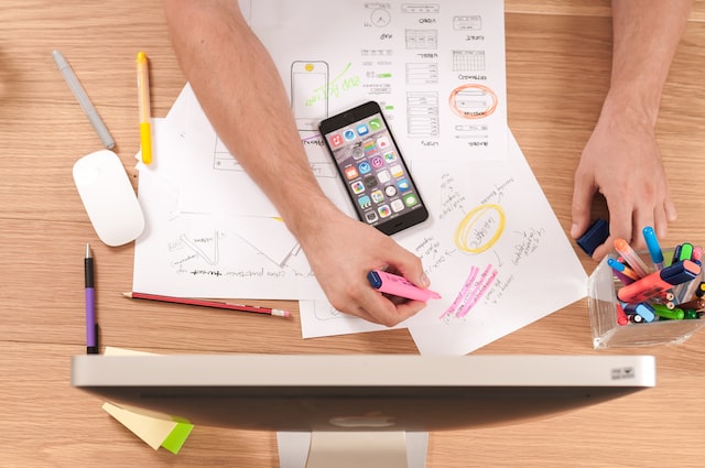 Overhead shot of someone's arms working on a desk with paper, a phone, and a desktop computer